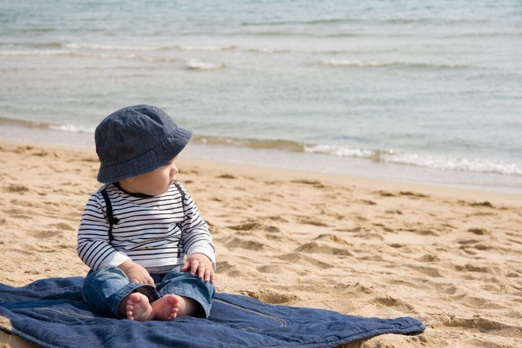 Young baby sitting on the beach on a blue blanket wearing a blue sun hat